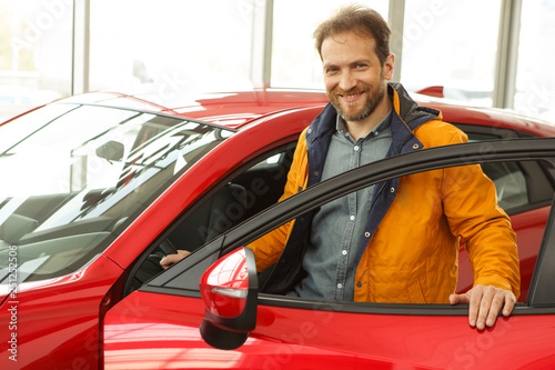 Handsome red haired male customer standing with opened door, near vehicle, smiling and looking at camera. Man enjoying comfortable red car, want buy new automobile in modern car dealership.