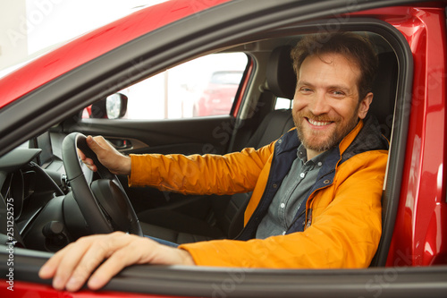 Red haired man enjoying comfortable seats of new red car. Male want buy new automobile in modern car dealership, sitting in vehicle, smiling and looking at camera. Concept of transportation. © Nestor