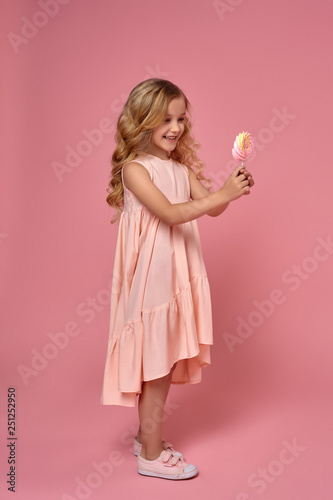 Little girl with a blond curly hair, in a pink dress is posing with a candy