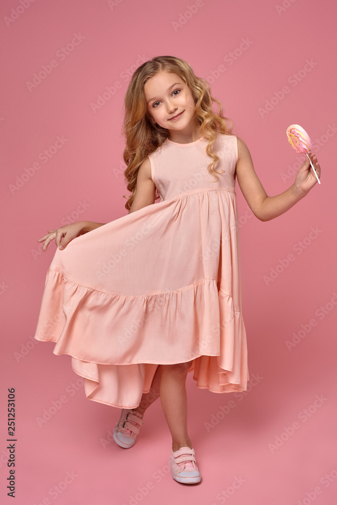 Little girl with a blond curly hair, in a pink dress is posing with a candy Little girl with a blond curly hair, in a pink dress is posing with a candy