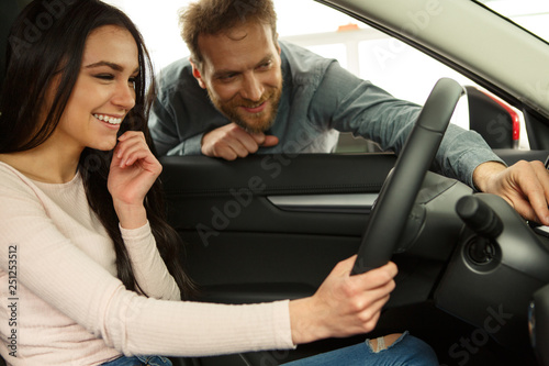 Side view of young beautiful woman sitting inside car and holding hand on steering wheel. She smiling and talking with manager of car dealership. Car agent representing inside of automobile.