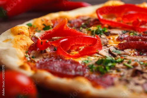 Close up Italian pizza with melted cheese, sausage, ham, salami, mushrooms, pepper and fresh herbs on a brown table decorated by mushrooms, red sweet pepper and cherry tomatoes