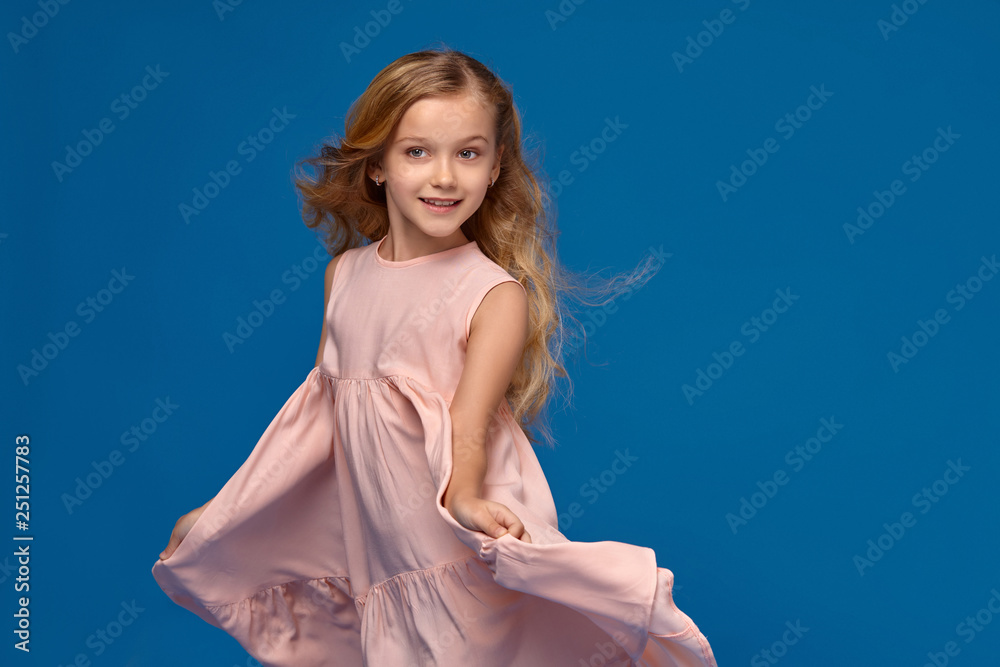 Fashionable little girl in a pink dress is posing on a blue background.