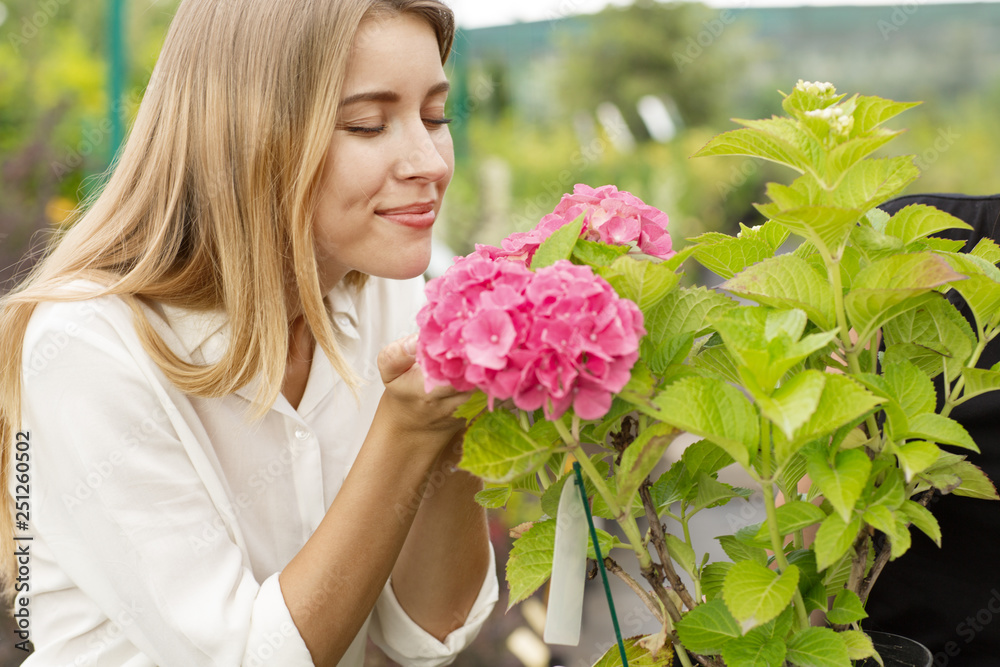 Attractive blonde with eyes closed enjoying scent of blooming flower in greenhouse. Female customer in white shirt smiling and keeping pink hydrangea in hands. Concept of pleasure. Attractive blonde with eyes closed enjoying scent of blooming flower in greenhouse. Female customer in white shirt smiling and keeping pink hydrangea in hands. Concept of pleasure.