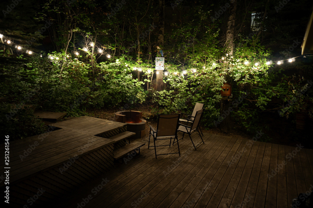 Patio lights strung through trees and a group of chairs sit around a fire pit on a wooden deck 