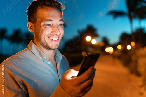 Man smiling looking at phone outside on city street at night texing online using smartphone. Young male face lit by screen light using mobile cellphone outdoors in the dark in summer. photo