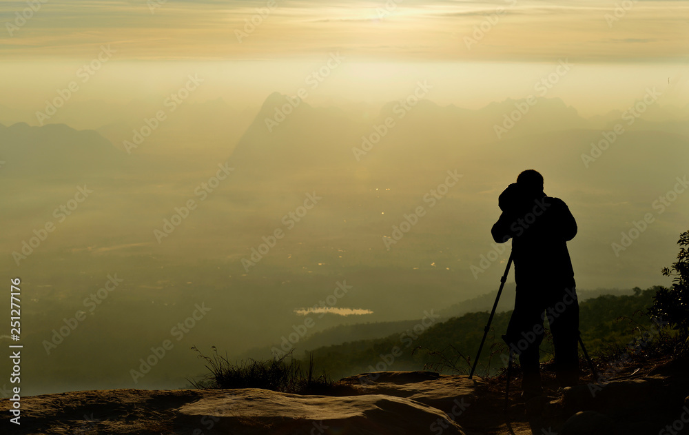 A photographer silhouette (on a cliff) is taking an image of mountain range in a morning