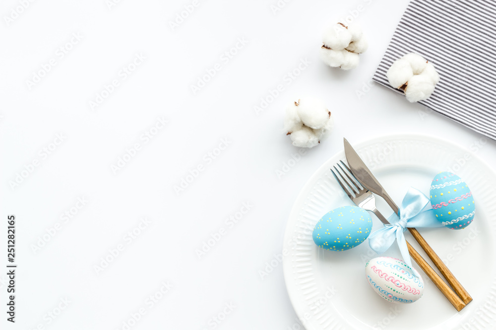 Fototapeta premium Easter table decoration in blue colors. Plate, cutlery, painted eggs and dry cotton flowers, tablecloth on white background top view space for text