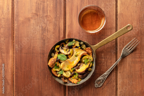 A photo of a rustic mushrooms and olives saute in a cast iron pan, with a glass of white wine, shot from above on a dark rustic background with copy space