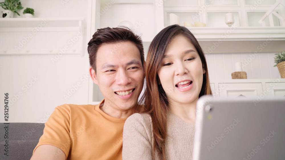 Fototapeta premium Asian couple using tablet VIDEO Call with friend in living room at home, sweet couple enjoy love moment while lying on the sofa when relaxed at home. Lifestyle couple relax at home concept.