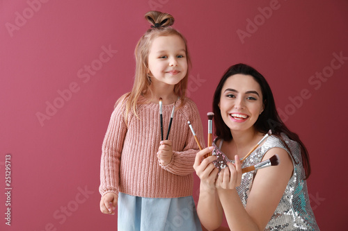 Portrait of cute little girl and her mother with makeup brushes on color background