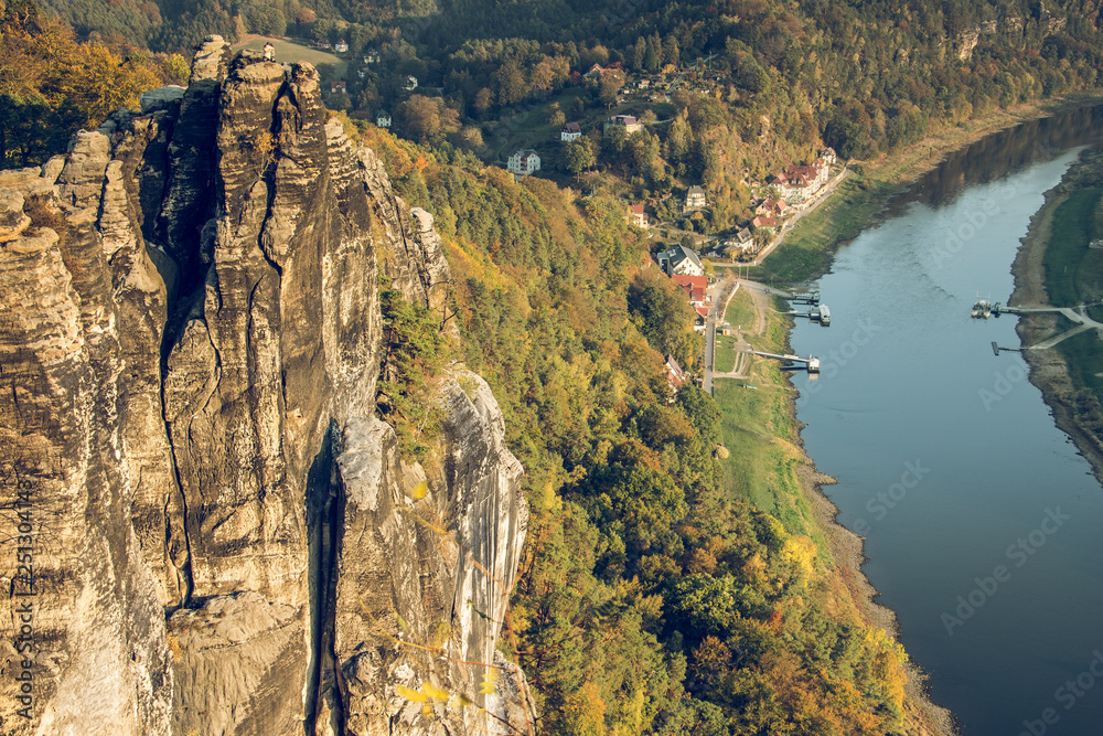 Rock formation in Saxon Switzerland. In the valley runs the river Elbe with ferry and buildings from Rathen. Forest in the evening sun in autumn.
