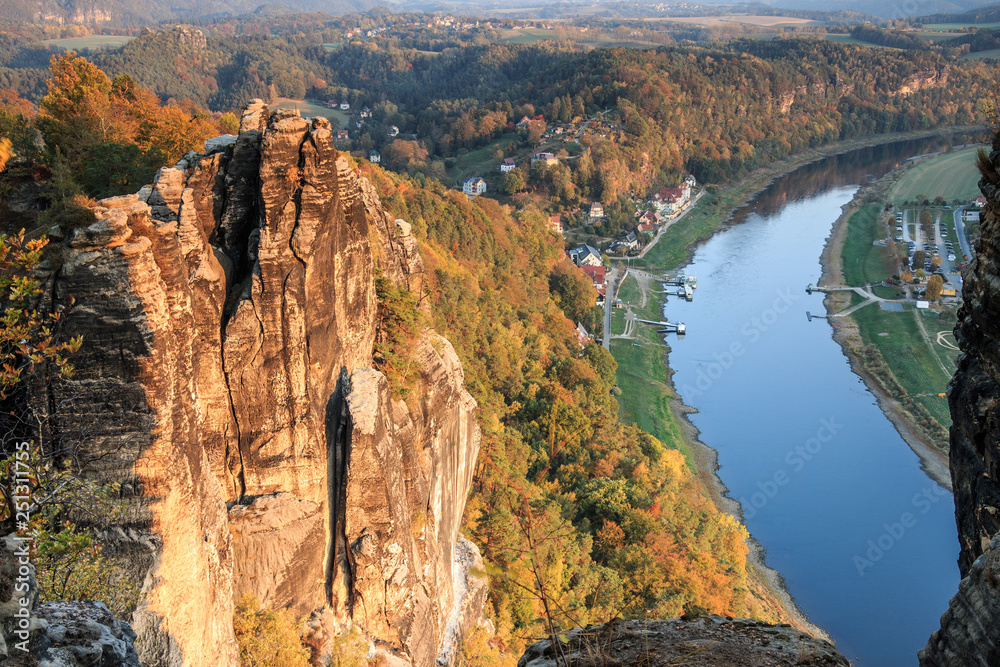 Saxon Switzerland National Park in the Elbe Sandstone Mountains. Rock formation and trees illuminated by evening sun in autumn. In the background forests and houses, in the valley river of the Elbe