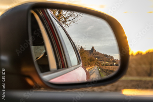 Close up of a car side mirror reflecting a ruiny castle and a road towards it during evening sunset, Trosky castle, Czech Bohemian paradise, Czech Republic