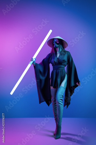 Concept on cosmic cosplay. Сontemporary portrait a young athletic woman in traditional Japanese black kimono, an Asian hat and highboots is holding a lightsaber and posing on neon blue-pink background