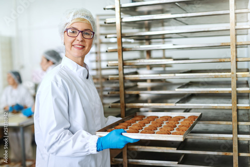 Smiling blonde female employee with eyeglasses and in sterile uniform putting tray with delicious cookies in rack. photo