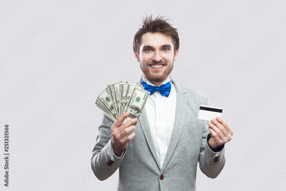 Handsome happy young bearded man in gray suit and blue bow tie standing and holding bank credit card and many cash dollars with toothy smile face. Indoor, isolated, studio shot, grey background Handsome happy young bearded man in gray suit and blue bow tie standing and holding bank credit card and many cash dollars with toothy smile face. Indoor, isolated, studio shot, grey background