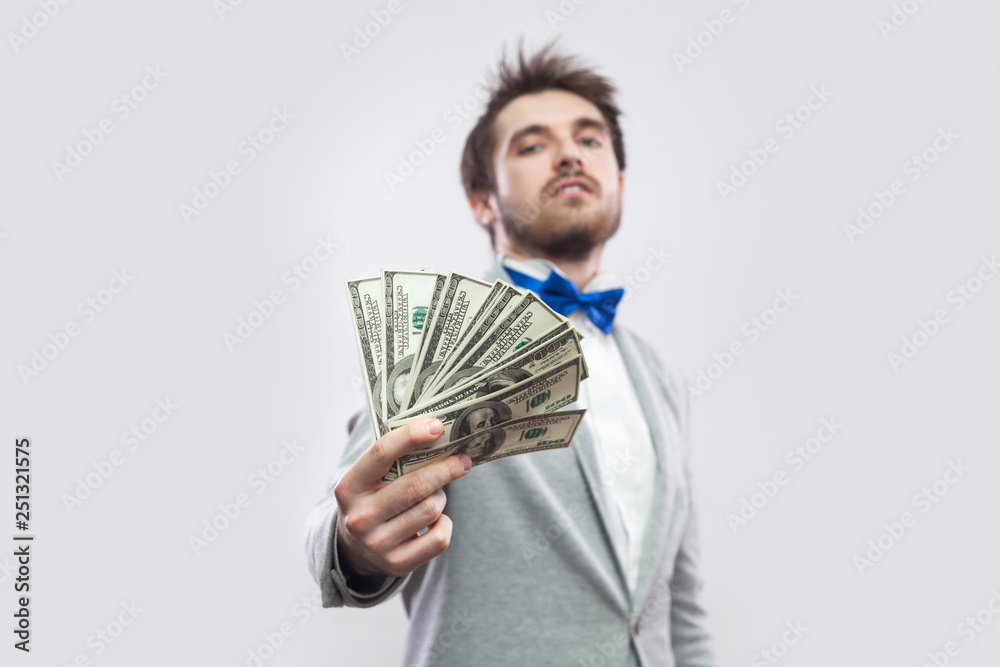 Take it! Portrait of confident attractive young businessman in gray coat and blue bow tie standing, proudly with head up and giving you many dollars. Indoor, studio shot, isolated, gray background. Take it! Portrait of confident attractive young businessman in gray coat and blue bow tie standing, proudly with head up and giving you many dollars. Indoor, studio shot, isolated, gray background.