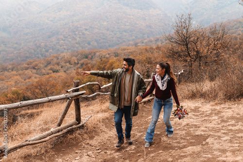 Couple in love walking in the nature at autumn and holding hands. Man pointing at view while woman holding bouquet in hand. In background forest and hills.