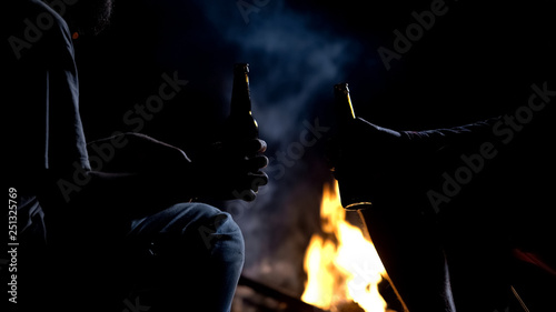 Men clinking beer bottles, nighttime camping, bonfire flaming, good company