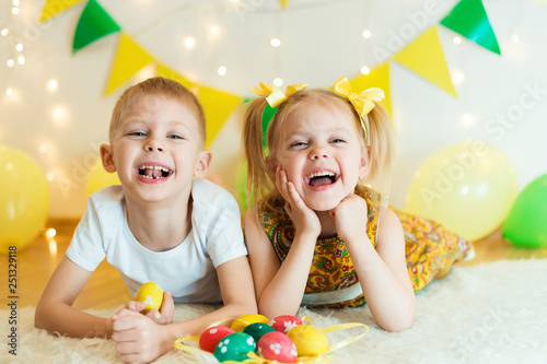blond, blue-eyed cute smiling children,boy in a white t-shirt and ponytail hair girl in yellow dress, brother and sister 4-5-6 years with Easter eggs in a room on the background of yellow decoration