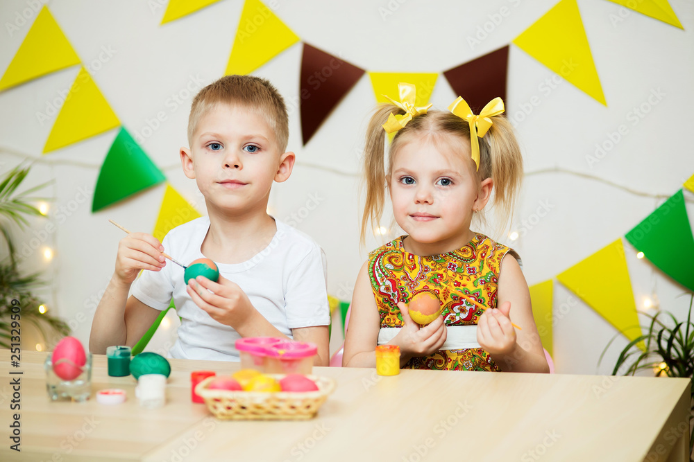 Fototapeta premium blond, blue-eyed cute smiling children, a boy in a white t-shirt and ponytail hair girl in a yellow dress, brother and sister 4-5-6 years painted Easter eggs in a room