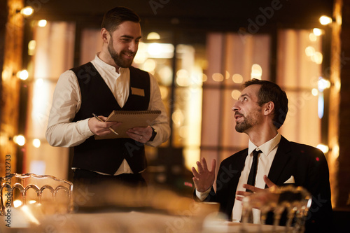 Portrait of mature gentleman ordering food in luxury restaurant with smiling waiter looking at him, copy space