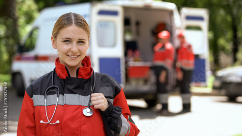 Female paramedic smiling into camera, ambulance crew blurred on background