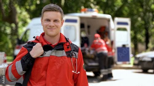 Male doctor smiling into camera, ambulance crew working, blurred on background photo