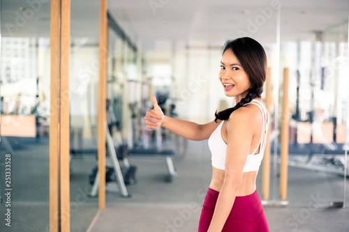 Young Asian fit and happy fitness female instructor giving thumbs up in professional sportswear inside gym background