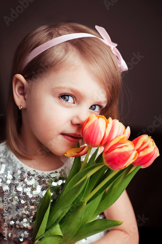 cute little blonde girl of three years with peach tulips close-up, smiling, look into the camera, vertical photo