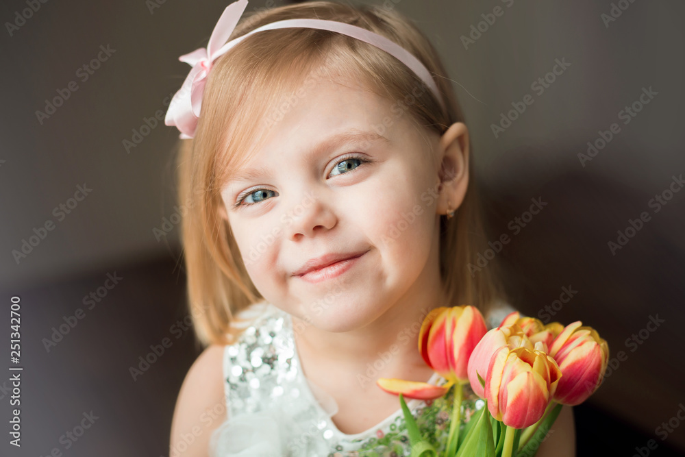 cute little blonde girl of three years with peach tulips close up, looking at the camera and smiling, her hair on her shoulders
