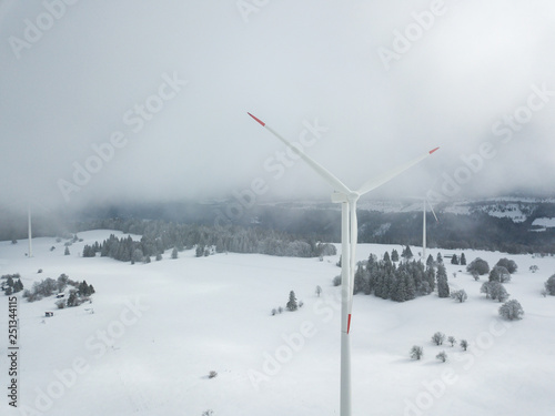 Aerial view of wind turbine in snow covered landscape in Swizterland. Tall pylon in fog, fir trees in the background. © Mario