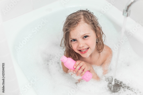 little girl 6 years old (baby) sitting in the bathroom in the water with fluffy foam and a shampoo bottle without tears in her hands