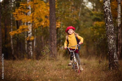 cheerful blonde babe 6 years old in a red hat yellow sweater in autumn riding a bike in the park or in the woods