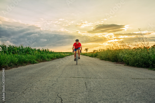 The cyclist in red blue form rides along fields of sunflowers. In the background sunset sky. Goes to camera.