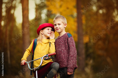 happy children babe 6 years old in red bloom and her brother, a boy of 7 years riding a bicycle in the park or in the woods in the golden autumn