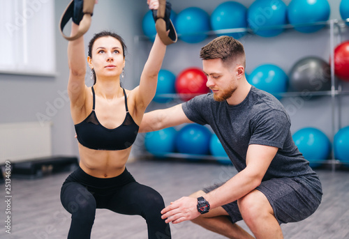 Muscular beardy trainer helping attractive female to practice with TRX in the fitness center. Sporty woman doing exercises with personal instructor at gym. Close-up