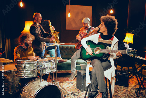 Mixed race woman playing acoustic guitar and sitting with legs crossed in home studio. In background the rest of the band playing drums, saxophone and bass guitar.