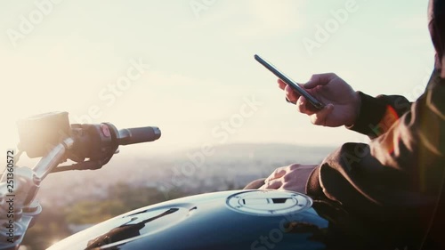 Motobiker man using mobile phone or smartphone and cheking or chatting or texting or googling on social network like instagram or facebook feed on sunrice or sunset outdoors conected to internet 4g photo