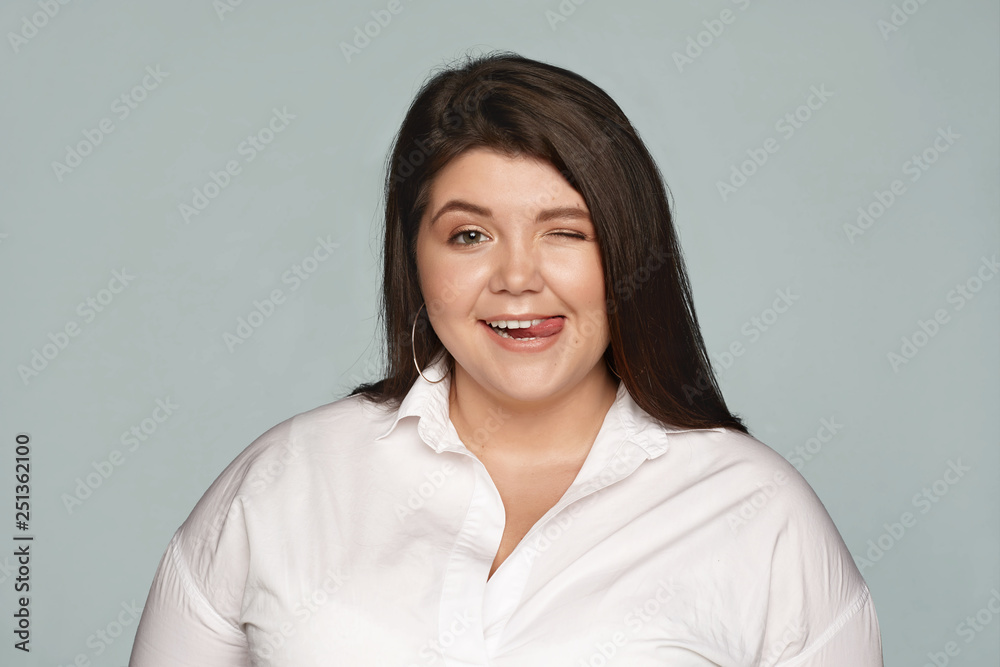 Isolated studio image of cheerful joyful chubby overweight female in formal white shirt having fun at office during break, winking and sticking out tongue at camera, flirting or teasing you Isolated studio image of cheerful joyful chubby overweight female in formal white shirt having fun at office during break, winking and sticking out tongue at camera, flirting or teasing you