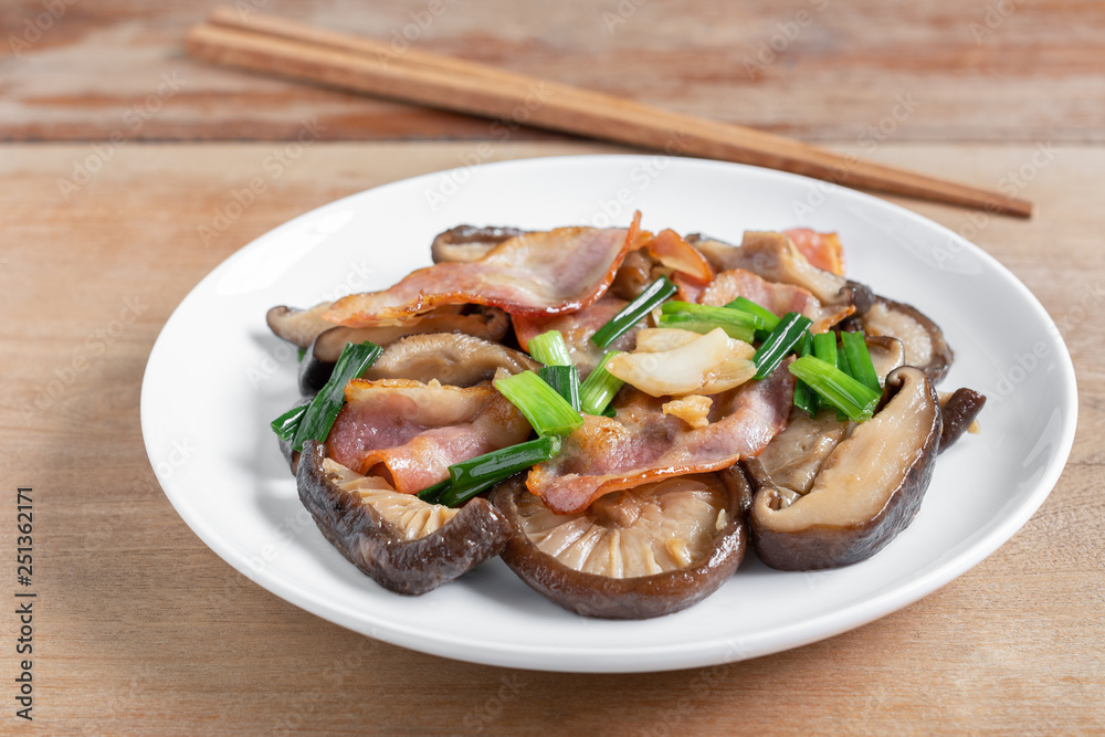 stir fried shiitake mushroom and bacon with soy sauce in a ceramic dish on wooden table, close up. homemade style food concept.