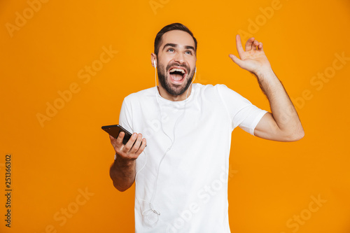 Photo of brunette man 30s listening to music using earphones and mobile phone, isolated over yellow background