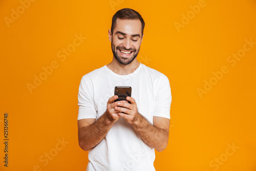Photo of joyful man 30s in casual wear smiling and holding smartphone, isolated over yellow background
