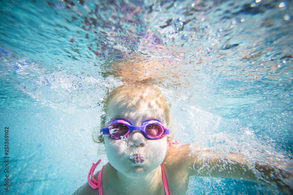 Wide angle underwater photo of a toddler girl swimming in a big swimming pool with goggles and a pink bikini Wide angle underwater photo of a toddler girl swimming in a big swimming pool with goggles and a pink bikini