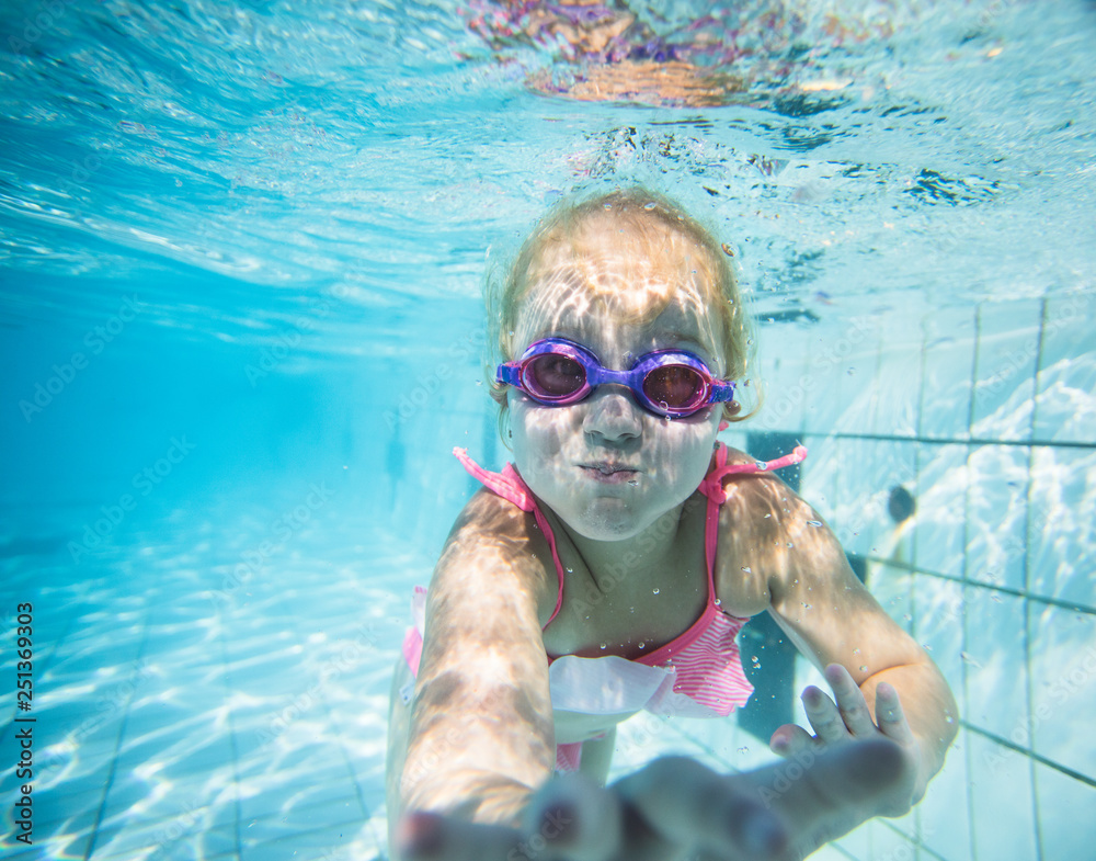 Fototapeta premium Wide angle underwater photo of a toddler girl swimming in a big swimming pool with goggles and a pink bikini