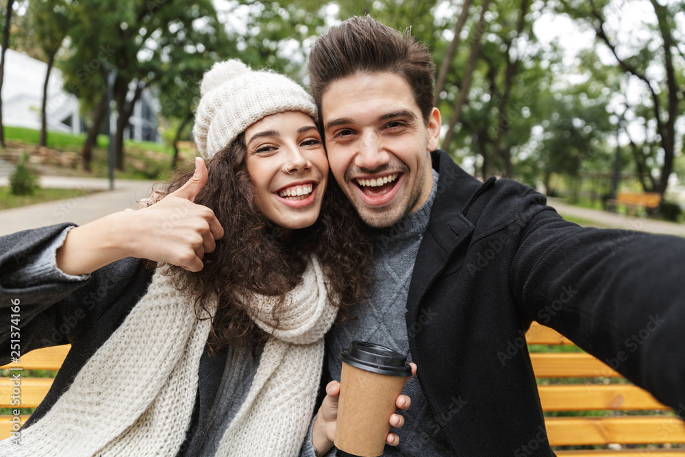 Fototapeta premium Happy young loving couple drinking tea or coffee outdoors in park talking with each other take a selfie by camera.