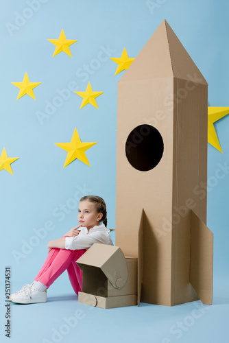 Dreamy kid with helmet sitting near cardboard rocket on blue starry background