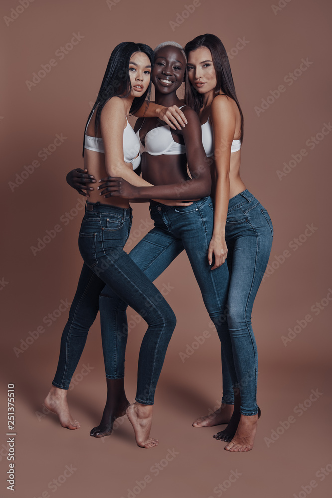 Looking gorgeous! Full length of three attractive young women looking at camera and smiling while standing against brown background Looking gorgeous! Full length of three attractive young women looking at camera and smiling while standing against brown background