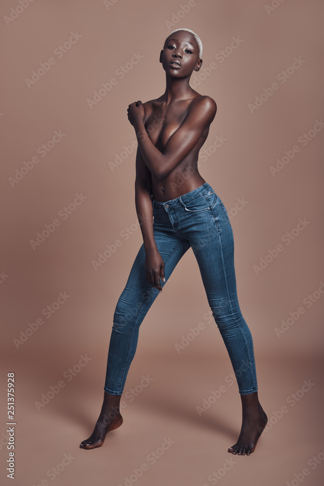Free and confident. Full length of attractive young African woman looking at camera and covering breast with hand while standing against brown background Free and confident. Full length of attractive young African woman looking at camera and covering breast with hand while standing against brown background